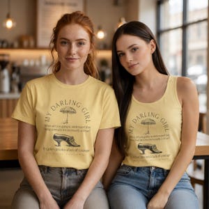 May include: A pale yellow short-sleeved t-shirt and a matching pale yellow tank top, both featuring a black graphic of an umbrella and a shoe with the text: My Darling Girl, When are you going to understand that being normal is not necessarily a virtue? It rather denotes a lack of courage.