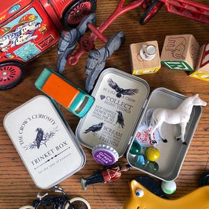 Puede incluir: Una colecci&oacute;n de juguetes y baratijas vintage, que incluye un cami&oacute;n de bomberos rojo, una motocicleta de juguete y varias cajas de metal peque&ntilde;as. Las cajas presentan ilustraciones de cuervos y texto, una de ellas etiquetada como "The Crow's Curiosity Trinket Box."