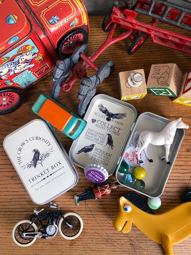 Puede incluir: Una colecci&oacute;n de juguetes y baratijas vintage, que incluye un cami&oacute;n de bomberos rojo, una motocicleta de juguete y varias cajas de metal peque&ntilde;as. Las cajas presentan ilustraciones de cuervos y texto, una de ellas etiquetada como "The Crow's Curiosity Trinket Box."