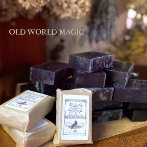 Op de afbeelding: Een display van donkerpaarse, vierkante zwarte zeepblokken, sommige verpakt in bruin papier met labels. De labels tonen een ravenillustratie en de tekst "Spellbound Black Soap" en "Old World Magic".
