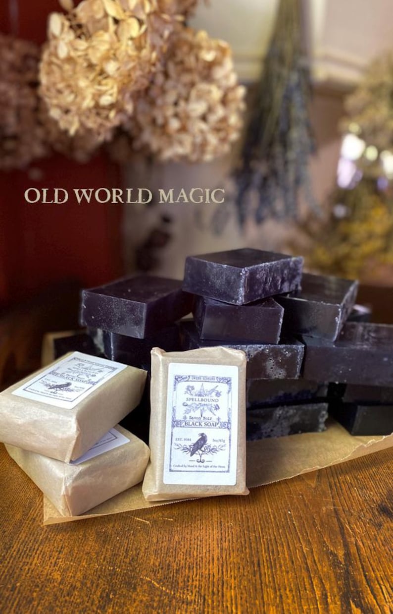 Op de afbeelding: Een display van donkerpaarse, vierkante zwarte zeepblokken, sommige verpakt in bruin papier met labels. De labels tonen een ravenillustratie en de tekst "Spellbound Black Soap" en "Old World Magic".