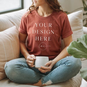 May include: A woman wearing a rust-colored t-shirt with the text "YOUR DESIGN HERE" in white. She is sitting on a couch with a white mug in her hands.