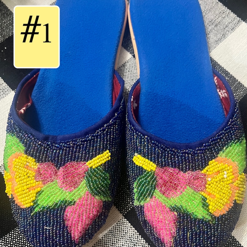 以下が含まれることがあります： 青いビーズのスリッパのペア、花の刺繍入り。 スリッパは青い内側で、黄色、ピンク、緑のビーズの花で飾られています。 テキスト「#1」は、左上隅の黄色の長方形にあります。
