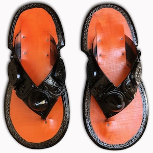 May include: A pair of black leather flip-flops with orange soles. The straps have a decorative design and a black button.
