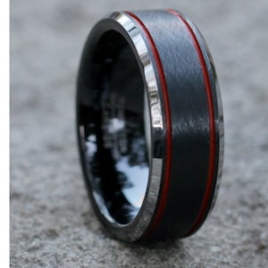 May include: Close-up of a black ring with a brushed metal finish. The ring features a red stripe in the centre and a silver-coloured edge. This ring is a piece of jewellery.