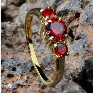 Pode incluir: Um anel de ouro com três pedras preciosas vermelhas. O anel está colocado sobre uma rocha marrom e áspera.