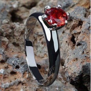 Pode incluir: Um anel de prata com uma gema vermelha engastada em garras. O anel está sobre uma superfície áspera e texturizada.