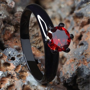 Könnte beinhalten: Ein schwarzer Ring mit einem großen, facettierten, roten Edelstein. Der Ring hat ein glattes, poliertes Band und ist vor einem strukturierten, felsigen Hintergrund platziert. Der Edelstein wird von vier schwarzen Zinken gehalten.