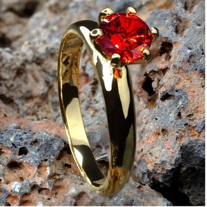 May include: A gold ring featuring a large, red gemstone. The ring is set against a backdrop of rough, brown rocks.