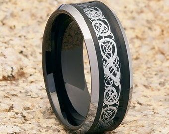 May include: A black ring with a silver Celtic knot design. The ring has a polished black band with a silver border. The ring is displayed on a light brown, textured surface. Jewelry.