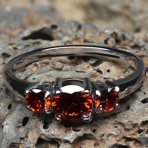 K&ouml;nnte beinhalten: Ein silberner Ring mit drei roten Edelsteinen, die in die Band gefasst sind. Der Ring liegt auf einer rauen, braunen, strukturierten Oberfl&auml;che.