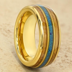 May include: A gold-colored ring with a wide band. The ring features a central inlay of blue-green material, flanked by wood-toned sections and accented with a gold-colored trim. The ring is designed with a polished finish.