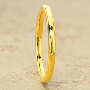 May include: A close-up shot of a gold ring. The ring is a solid band with a smooth, polished surface. The ring is a bright, shiny gold color. The background is a neutral, light brown color.