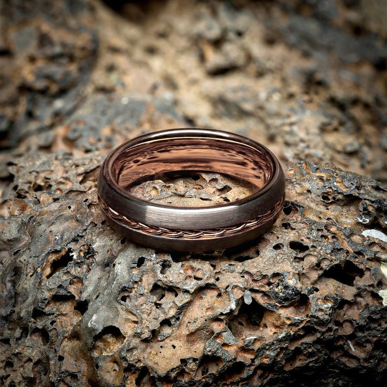 May include: A rose gold wedding band with a braided design. The band is sitting on a rough, textured surface.