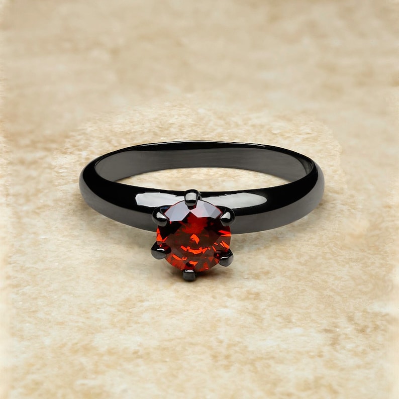 May include: A black band ring with a red gemstone in a prong setting.