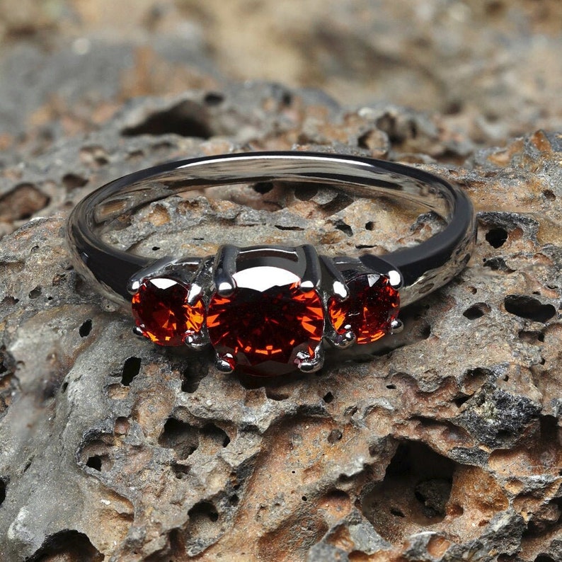 K&ouml;nnte beinhalten: Ein silberner Ring mit drei roten Edelsteinen, die in die Band gefasst sind. Der Ring liegt auf einer rauen, braunen, strukturierten Oberfl&auml;che.