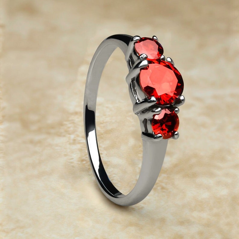 K&ouml;nnte beinhalten: Ein silberner Ring mit drei roten Edelsteinen. Der Ring hat eine schlichte Band und die Edelsteine sind in einer Krappenfassung gefasst.