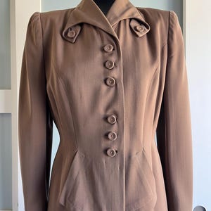 May include: A brown vintage jacket with a tailored fit, featuring a notched collar and decorative buttons. The jacket has long sleeves and a fitted waist, showcasing a classic, elegant design. The jacket is displayed on a black mannequin.