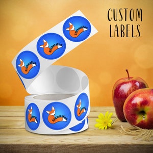 May include: Roll of round custom labels with a blue background and a cartoon fox with the text "StickersFox" in white. The labels are on a wooden surface with two red apples and a yellow flower.