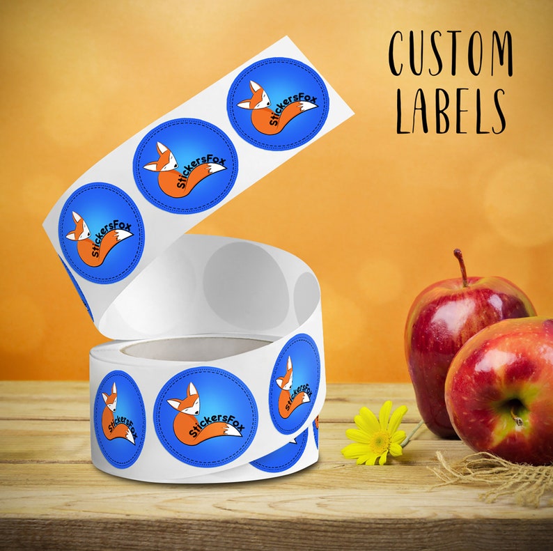 May include: Roll of round custom labels with a blue background and a cartoon fox with the text "StickersFox" in white. The labels are on a wooden surface with two red apples and a yellow flower.