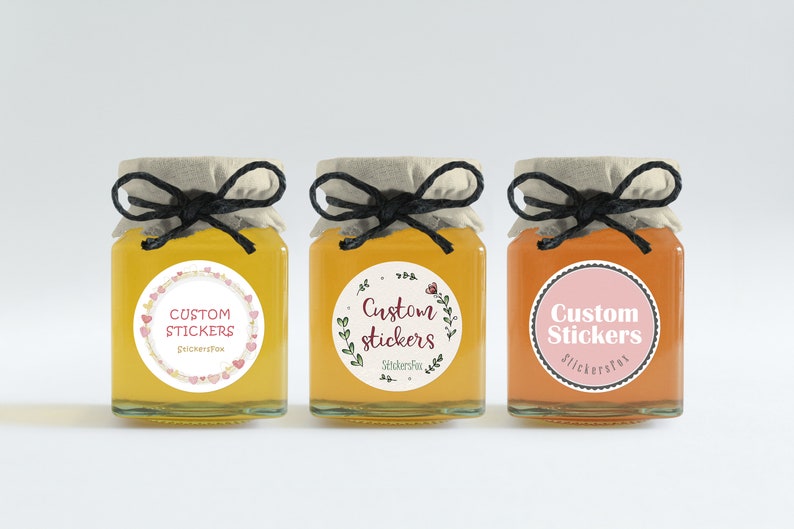 May include: Three glass jars with lids tied with black twine. Each jar has a round sticker with the text "Custom Stickers" and a different design. The first sticker has a pink and white heart design, the second has a green floral design, and the third has a pink and white polka dot design.