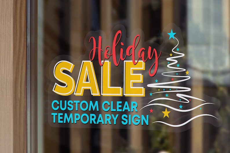 May include: A clear, custom temporary sign with the text "Holiday Sale" in yellow and red lettering. A stylized white Christmas tree with blue and yellow stars is on the right side of the sign.