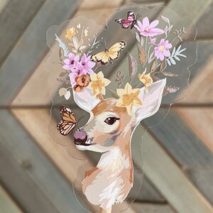 May include: A clear sticker with a fawn's head and neck, surrounded by a floral wreath with pink and yellow flowers and butterflies. The fawn has a butterfly on its nose.