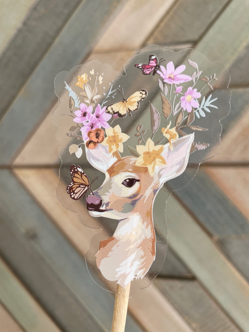 May include: A clear sticker with a fawn's head and neck, surrounded by a floral wreath with pink and yellow flowers and butterflies. The fawn has a butterfly on its nose.