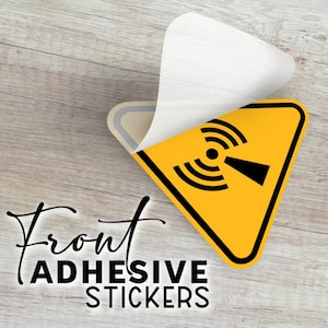 May include: Yellow and black triangular sticker with a radiation hazard symbol, partially peeled to reveal the adhesive backing. The text "Front ADHESIVE STICKERS" is displayed in a stylish font. The sticker is on a light wood grain surface.