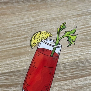 May include: A clear glass filled with a red liquid, garnished with a lime wedge and a sprig of celery. The glass is on a wooden stick.