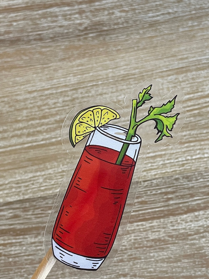 May include: A clear glass filled with a red liquid, garnished with a lime wedge and a sprig of celery. The glass is on a wooden stick.