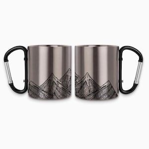 May include: Two stainless steel travel mugs with black carabiner handles. Each mug features a black and white mountain range design. The mugs are ideal for outdoor enthusiasts and camping.