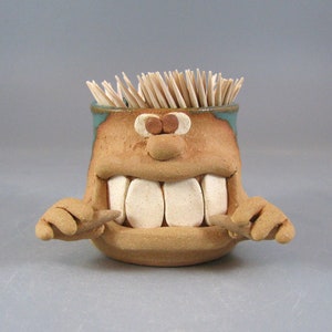 May include: A whimsical, handmade ceramic toothpick holder. The holder is shaped like a smiling face with large white teeth, brown eyes, and arms. The top rim is teal, and the holder is filled with wooden toothpicks.