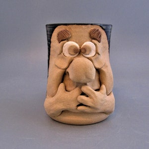 May include: A beige ceramic mug with a sculpted face design. The mug features a large nose, wide eyes, and hands clasped over the chest. The rim is dark blue. A unique and artistic piece for coffee or tea.