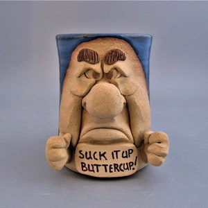 May include: A ceramic mug with a grumpy face design. The mug is tan with a blue rim and features a large nose, furrowed eyebrows, and the phrase "SUCK IT UP BUTTERCUP!" written on the front.