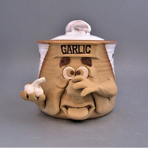 May include: A whimsical ceramic garlic keeper with a face design. The tan-colored container has a white lid and the word "GARLIC" in black. The container features a sculpted face with a garlic clove in one hand.