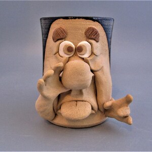 May include: A ceramic mug shaped like a cartoon face with a large nose, wide eyes, and outstretched arms. The mug is glazed in brown and blue.
