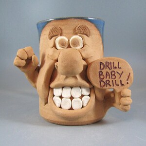 May include: A brown ceramic mug with a cartoon face, large nose, and white teeth. The mug is holding a sign that says "Drill Baby Drill!"