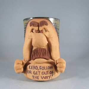 May include: A tan ceramic mug featuring a sculpted face with a prominent nose, thick eyebrows, and a dark brown mustache. The mug displays two raised fists and a plaque with the text, "LEAD, FOLLOW OR GET OUT OF THE WAY!" in dark lettering.