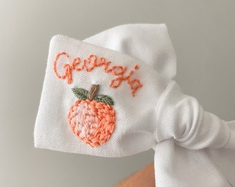Op de afbeelding: Witte stoffen strik met het woord "Georgia" geborduurd in roze draad en een perzik geborduurd in roze en groene draad.