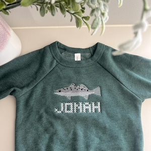 May include: A dark green sweatshirt with a fish design and the name "JONAH" embroidered in white. The fish is gray and white with black dots. The sweatshirt has a crew neck and long sleeves.