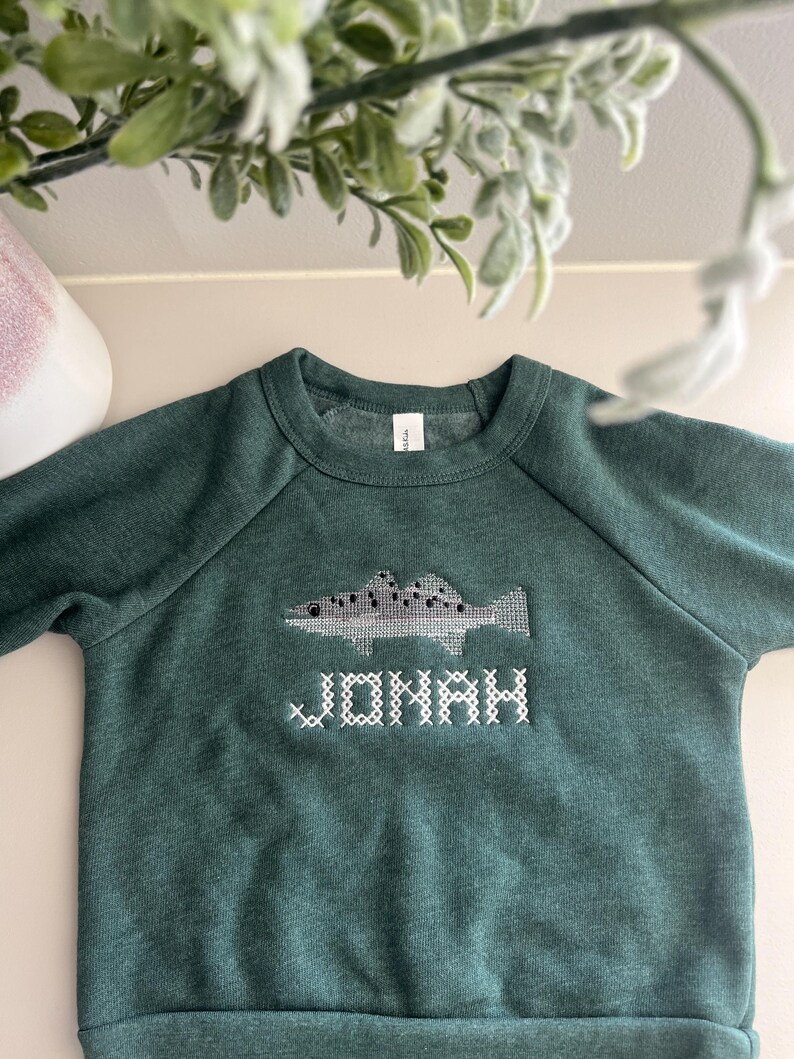 May include: A dark green sweatshirt with a fish design and the name "JONAH" embroidered in white. The fish is gray and white with black dots. The sweatshirt has a crew neck and long sleeves.