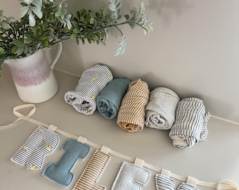 Op de afbeelding: Een stoffen banner met de naam RILEY, waarbij elke letter is gemaakt van verschillende texturen, waaronder blauwe, beige en zwart-wit gestreepte stof met kleine gele autootjes, vergezeld van vijf opgerolde stoffen artikelen.