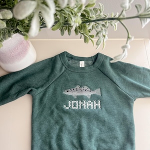 May include: A green sweatshirt with a fish design and the name "JONAH" embroidered in white. The sweatshirt has a crew neck and long sleeves. The fish design is in shades of gray and white.