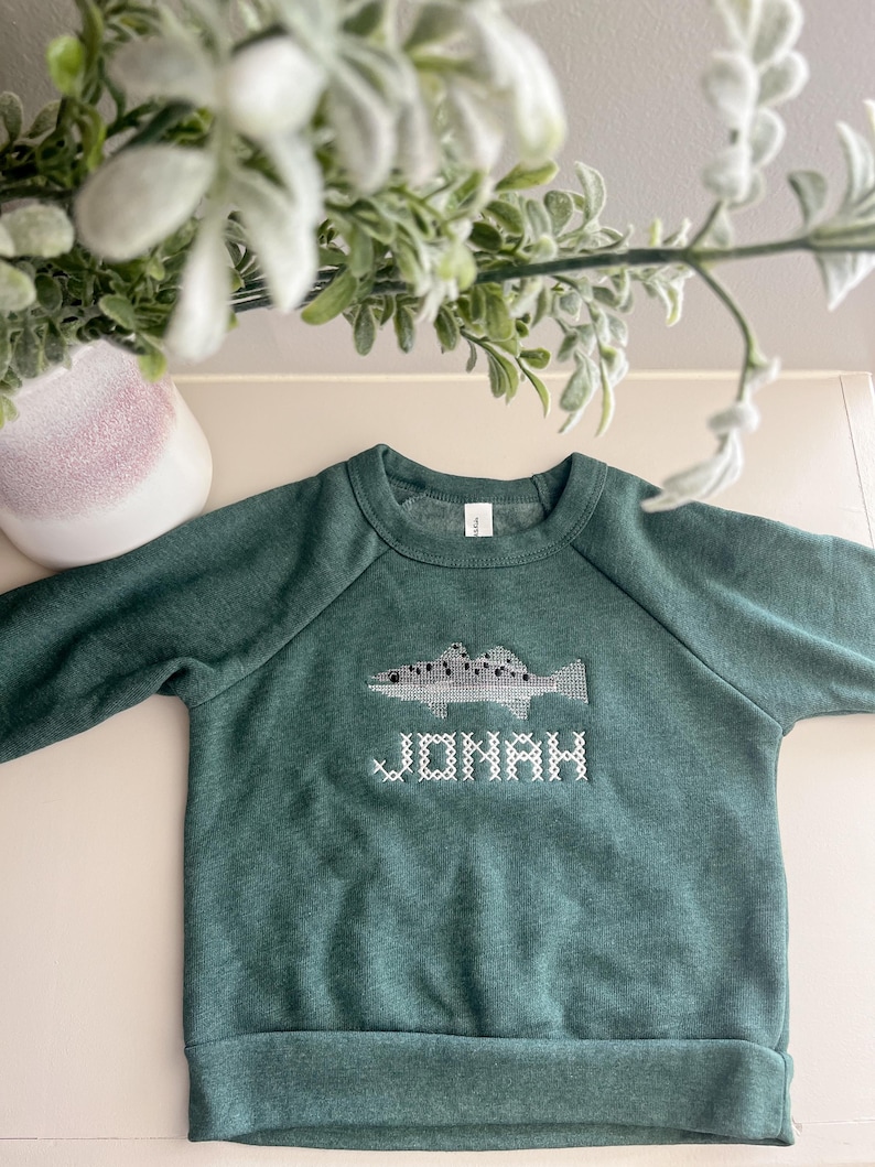 May include: A green sweatshirt with a fish design and the name "JONAH" embroidered in white. The sweatshirt has a crew neck and long sleeves. The fish design is in shades of gray and white.