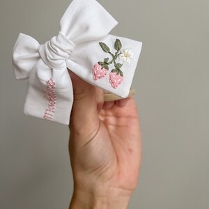 Op de afbeelding: Witte stoffen strik met geborduurde roze aardbeien, groene bladeren en een kleine witte bloem. Het woord "Maddie" is roze op de strik geborduurd. Een charmant haaraccessoire.