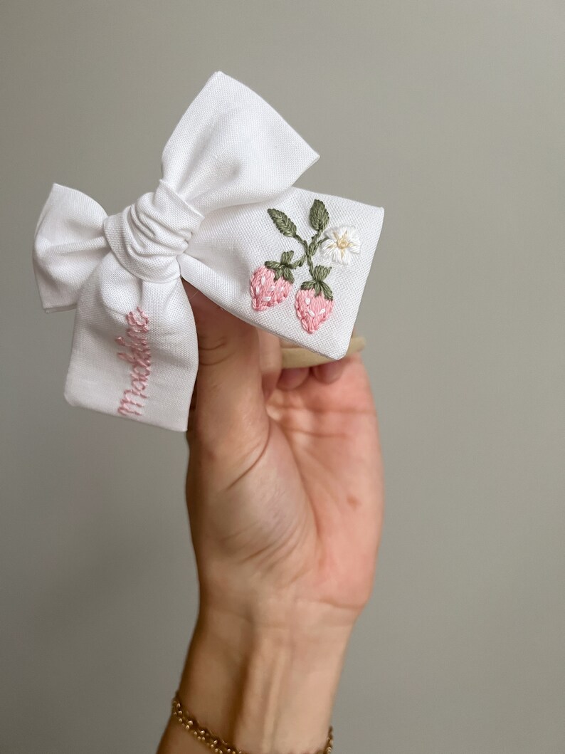 Op de afbeelding: Witte stoffen strik met geborduurde roze aardbeien, groene bladeren en een kleine witte bloem. Het woord "Maddie" is roze op de strik geborduurd. Een charmant haaraccessoire.