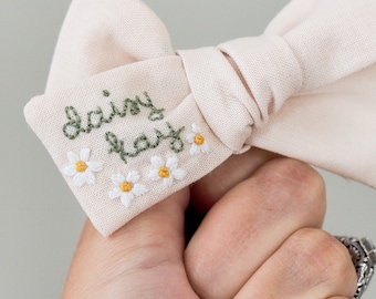 Op de afbeelding: Een lichtroze stoffen strik met de woorden "daisy farm" geborduurd in groene draad. De strik is versierd met vijf witte geborduurde madeliefjes.