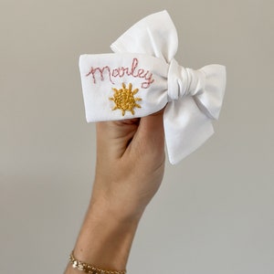 May include: White fabric bow with the name "Marley" embroidered in pink thread. A yellow sun design is embroidered below the name. The bow is held in a hand, with a gold bracelet visible.