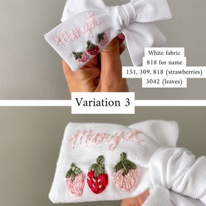 Op de afbeelding: Witte stoffen haarstrik met geborduurde naam en aardbeienontwerp. De naam is lichtroze en de aardbeien zijn in roze en rode tinten met groene bladeren. Variatie 3.
